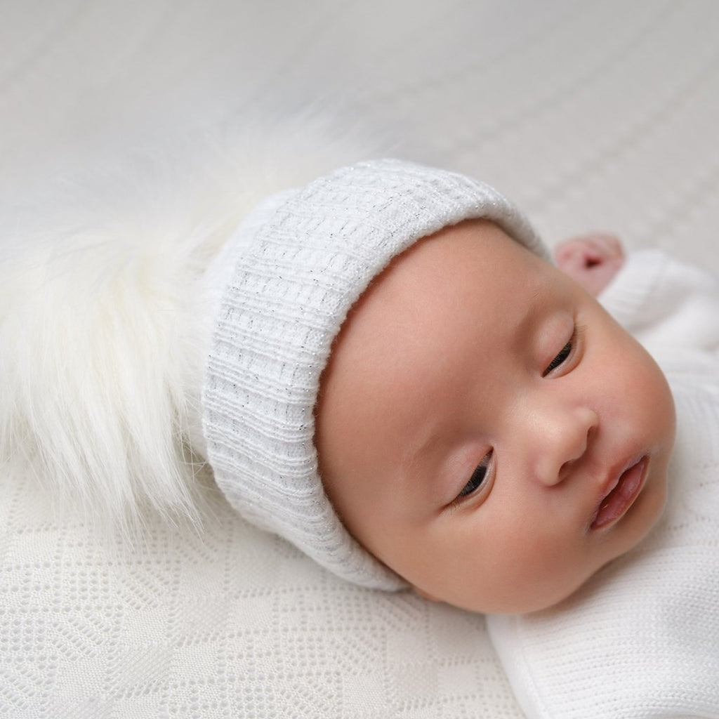 Baby on white blanket wearing the first size white and silver ribbed fur double pom hat