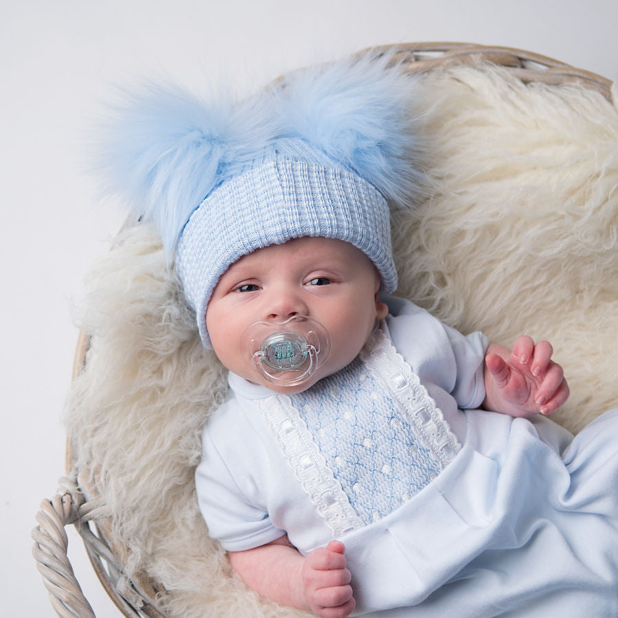 Baby with dummy wearing first size blue ribbed fur double pom pom hat