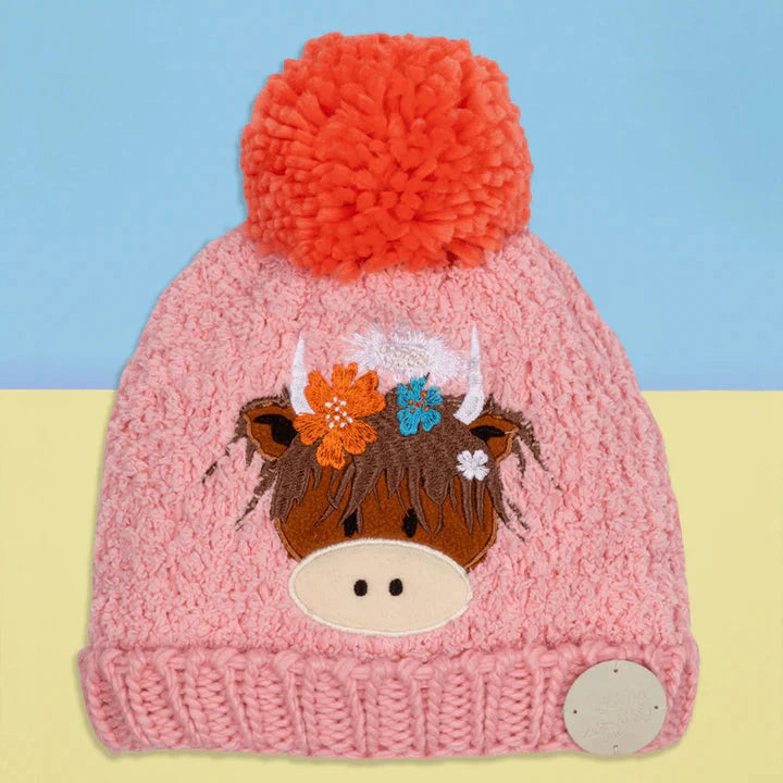 Pink knit hat with a cow design and orange pom-pom on a blue and yellow gradient background