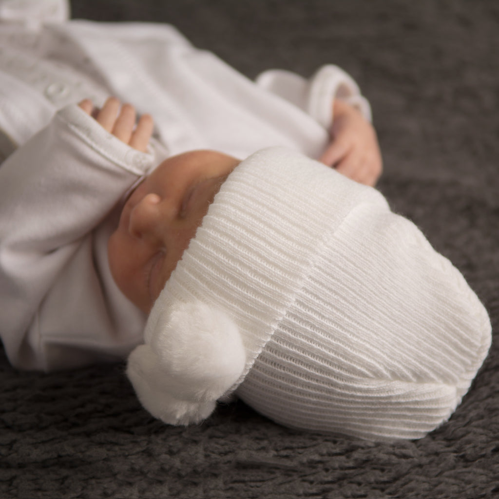 Baby on grey fur blanket wearing first size white hat with white pom pom