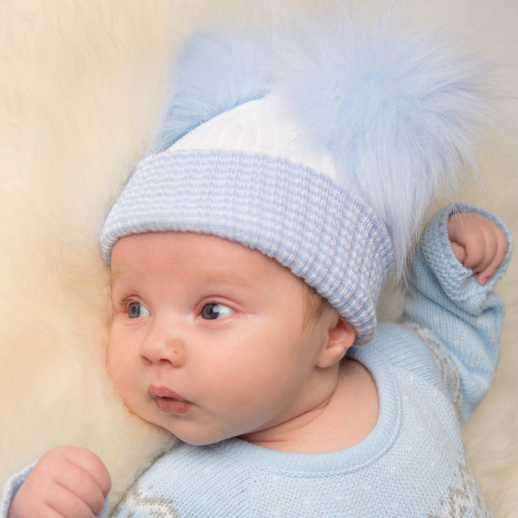 Baby on cream fur blanket wearing first size white and blue ribbed fur double pom hat