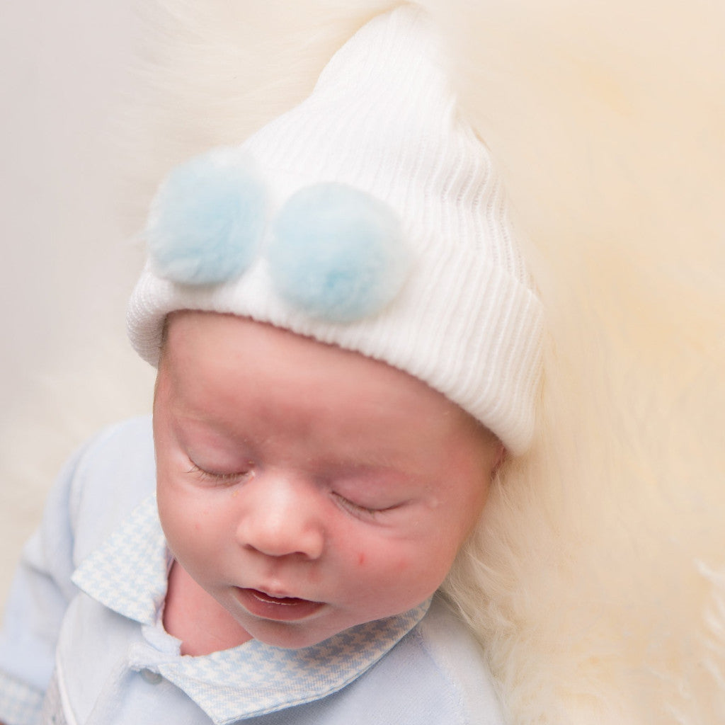 Baby wearing first size white hat with blue fur pom pom