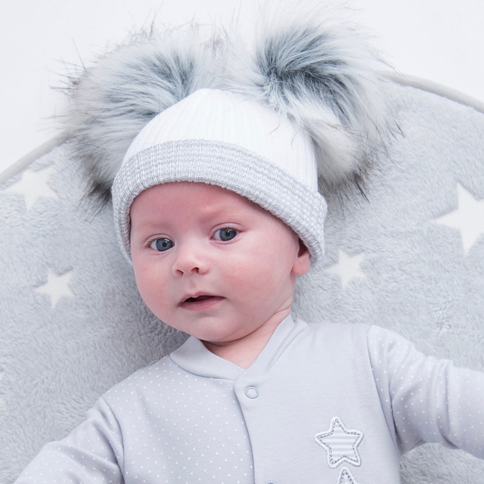 Baby on grey star rug wearing first size white and grey ribbed fur double pom hat