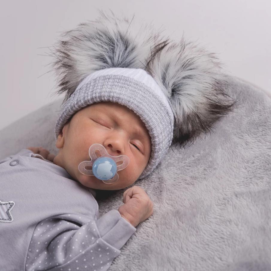 Sleeping baby with dummy on grey rug wearing the first size white and grey ribbed fur double pom hat