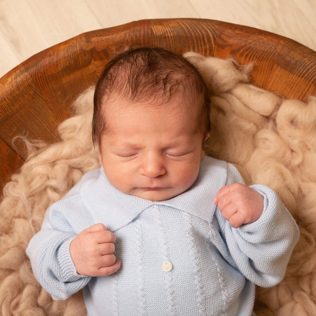 Close up of baby in wooden bowl on fur blanket wearing blue polo knitted jumper and trousers set