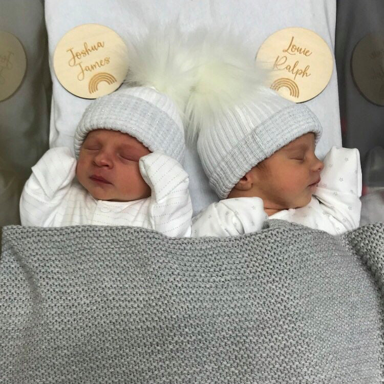 Two babies with name plaque's wearing the first size white and silver ribbed fur pom hats