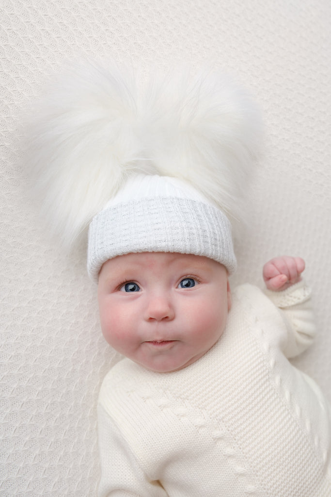 Baby with arm up on white blanket wearing First Size White And Silver Ribbed Fur Double Pom Hat
