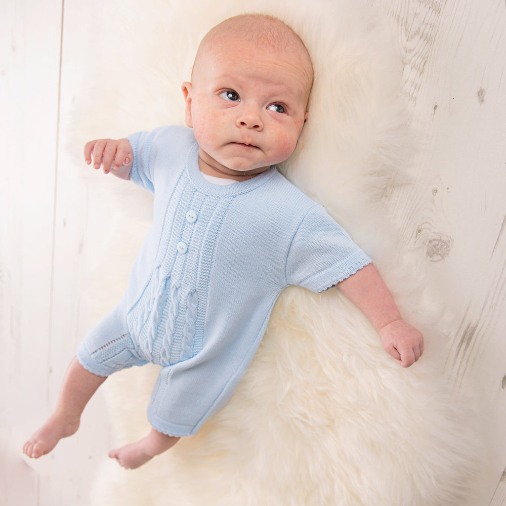 Baby looking up on fur blanket and wooden floor wearing blue cable knitted romper