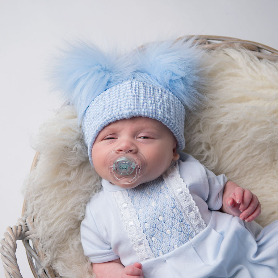 Baby in a basket on a fur blanket wearing first size blue ribbed fur double pom pom hat