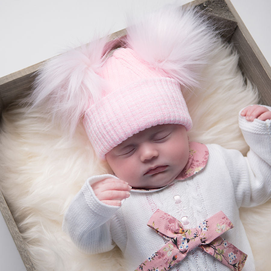 Sleeping baby in box on fur blanket wearing first size pink ribbed fur double pom hat