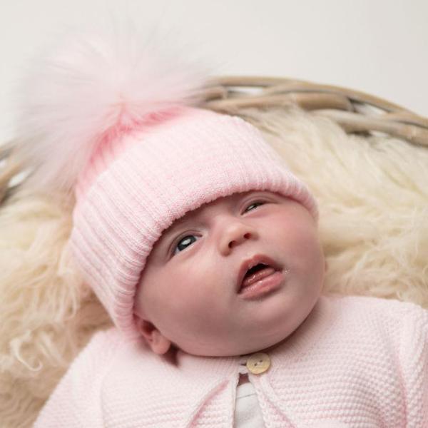Baby in basket on cream fur blanket wearing first size pink ribbed fur pom hat